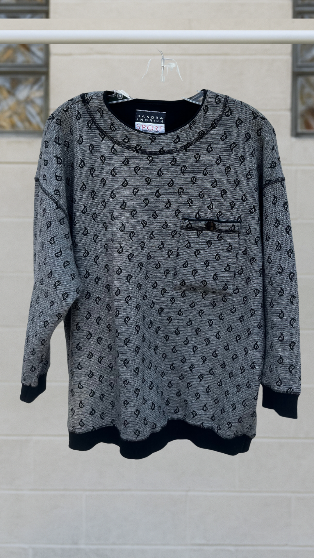 Grey paisley jacquard pullover with 3/4 sleeves, ribbed cuffs and hem, and a button-flap chest pocket; hanging on a hanger against a light block-glass wall.