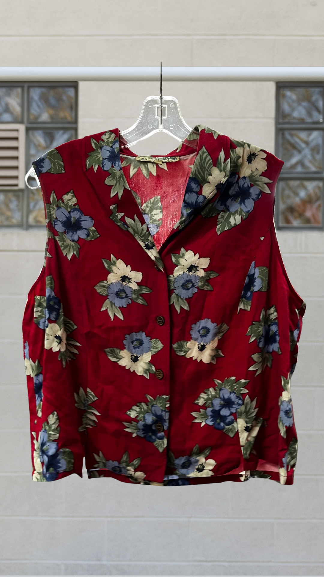 Sleeveless crimson rayon shirt with multi-color floral print, pointed collar, and wooden buttons, displayed front-on against light brick wall.