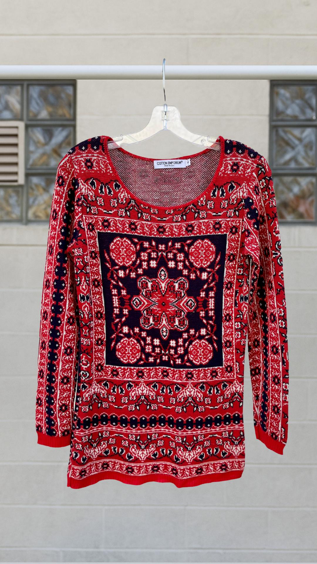 Red, navy, and white patterned long sleeve knit shirt with a central tapestry-inspired design, displayed on a clear hanger against a neutral background. Scoop neckline with thread detailing.