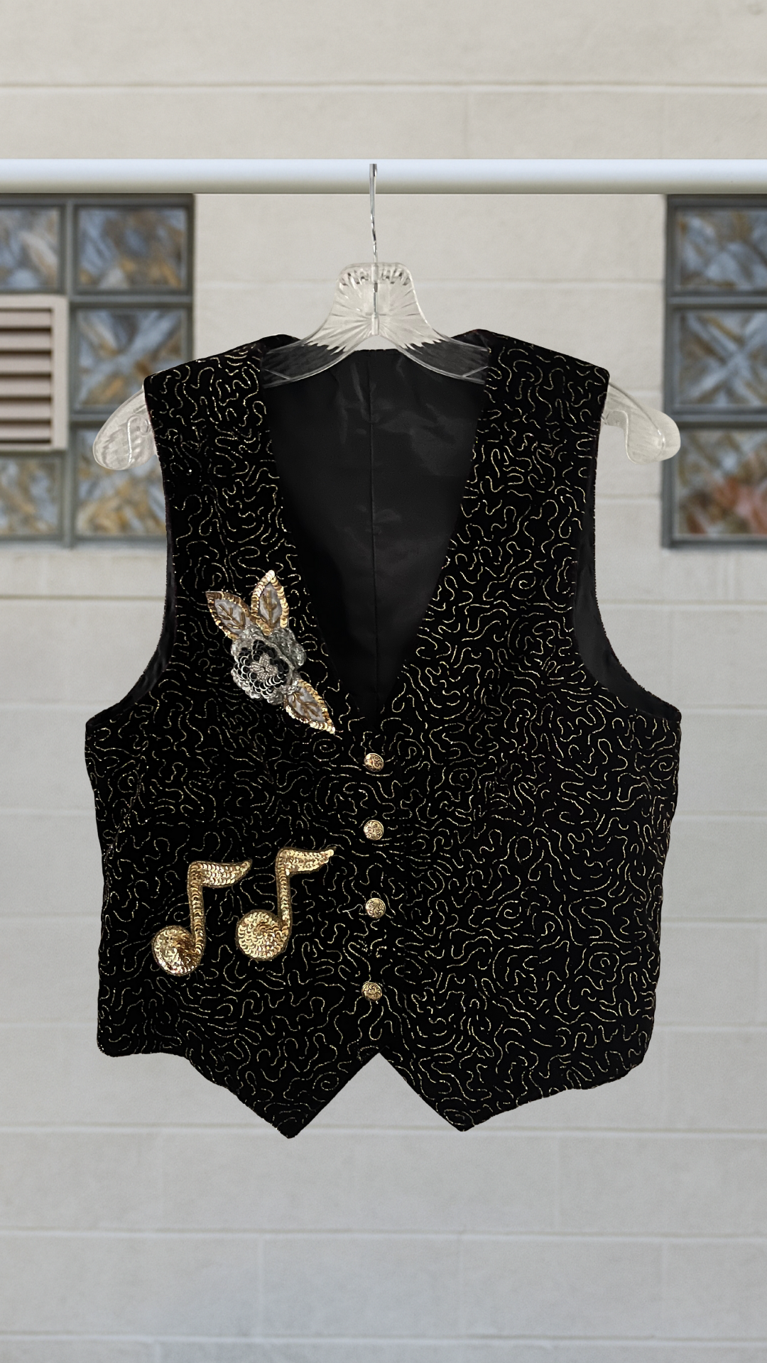 a black velvet vest covered in gold filigree and musical notes on a hanger against a white background