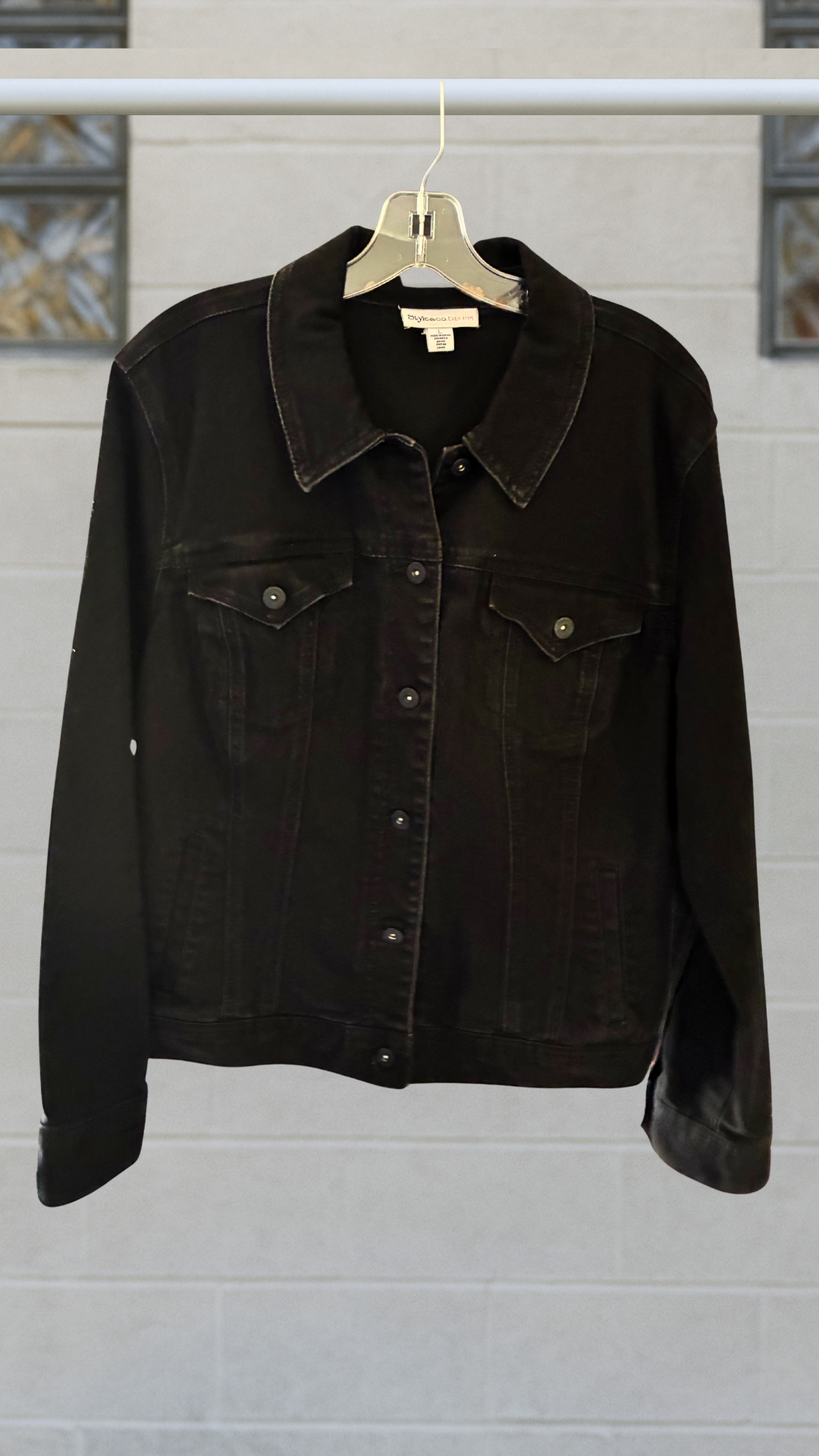 Black denim jacket with classic seaming, front button closure, and chest pockets; hanging on a hanger against a light block-glass wall.
