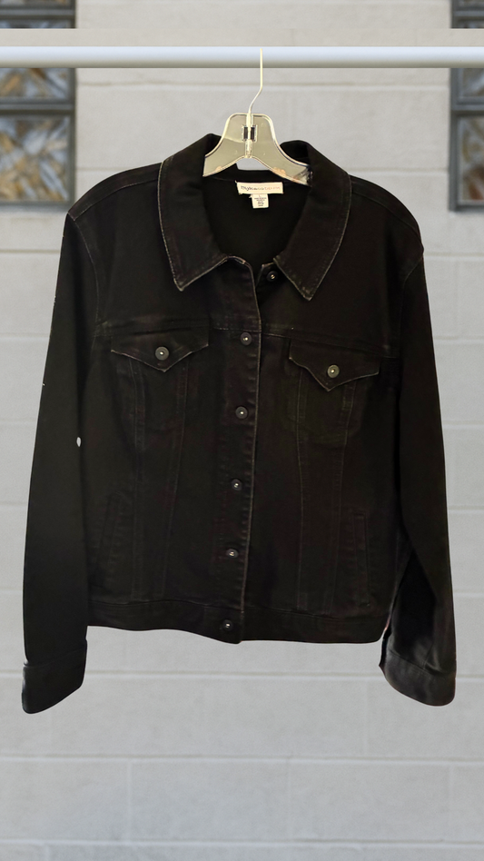 Black denim jacket with classic seaming, front button closure, and chest pockets; hanging on a hanger against a light block-glass wall.
