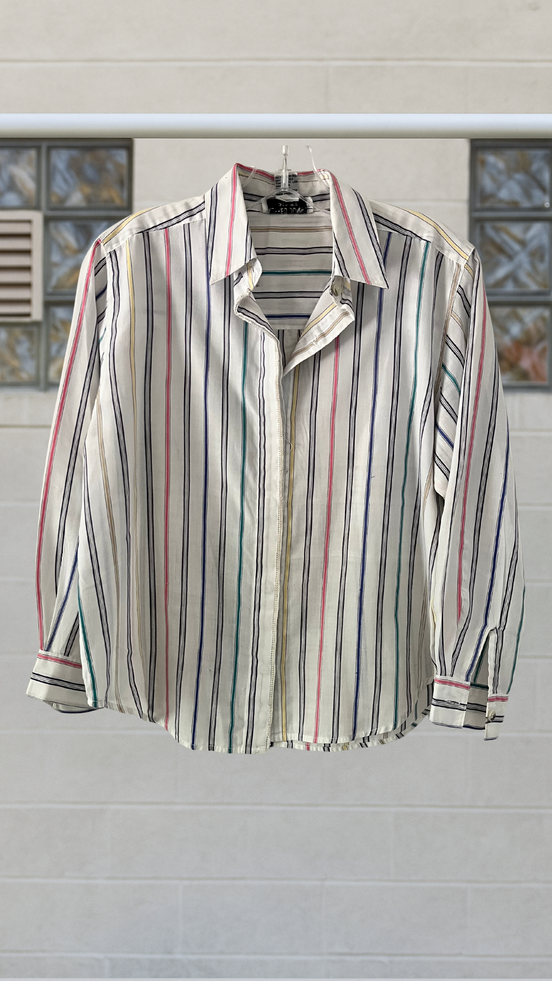 White button-down shirt with multicolor vertical stripes, long sleeves, and point collar, displayed front-on against light brick wall.
