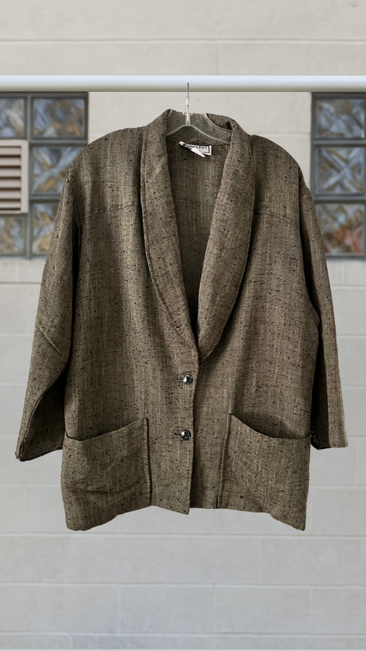 Soft earth‑tone woven blazer with lapel collar, two buttons, and large front pockets, hanging against a light wall.

