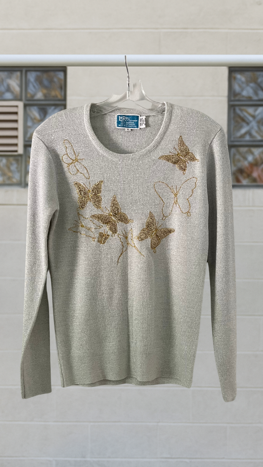 metallic sweater with butterfly designs on a hanger against a light-colored wall.