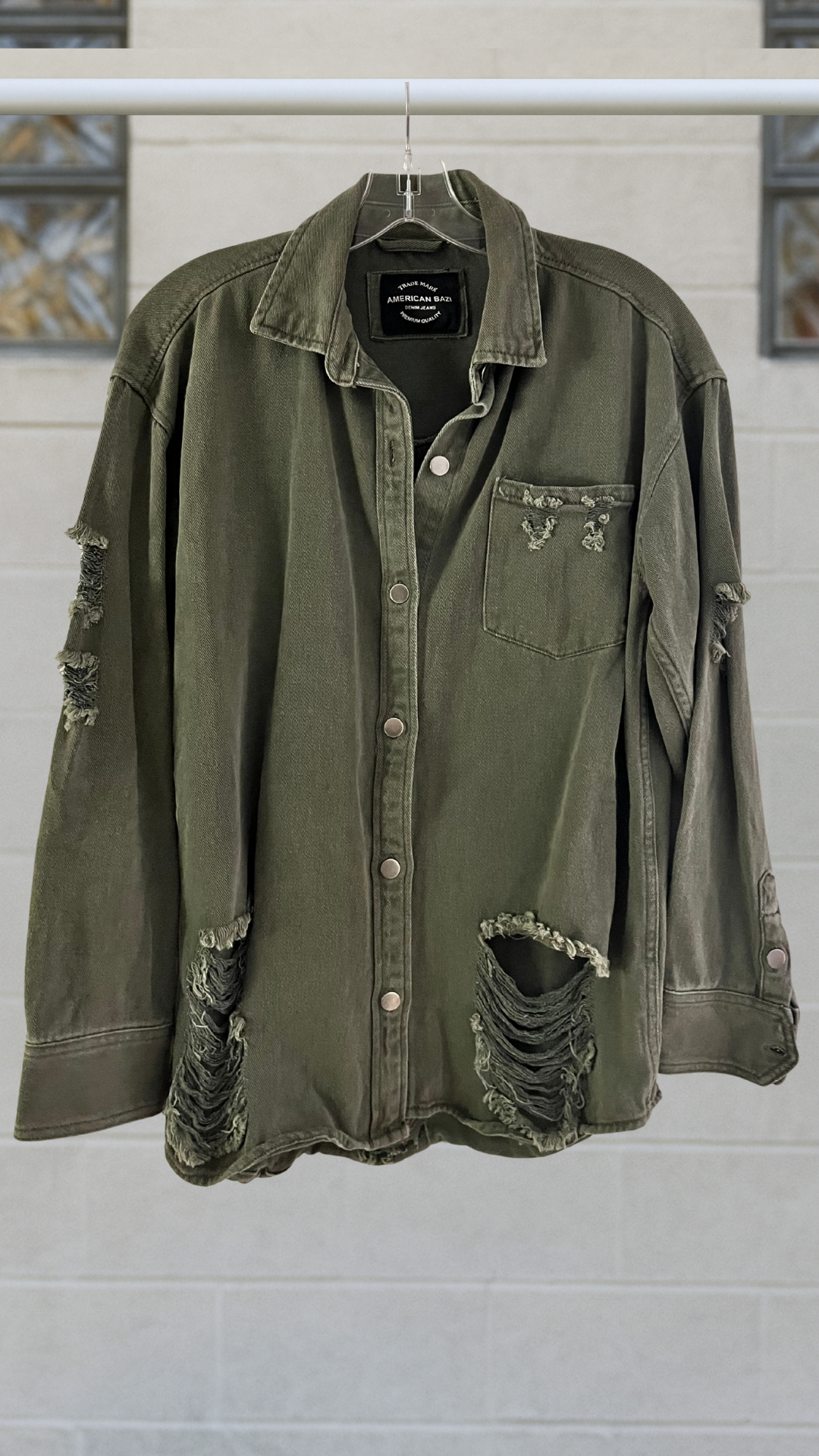 Olive green distressed denim snap-front shirt with long sleeves, chest pocket, and frayed holes; hanging on a hanger against a light block-glass wall.
