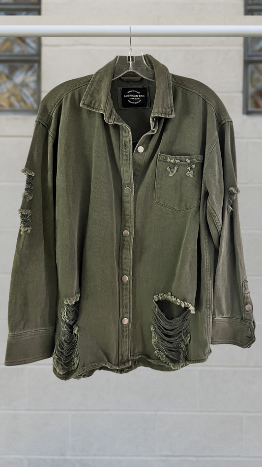 Olive green distressed denim snap-front shirt with long sleeves, chest pocket, and frayed holes; hanging on a hanger against a light block-glass wall.
