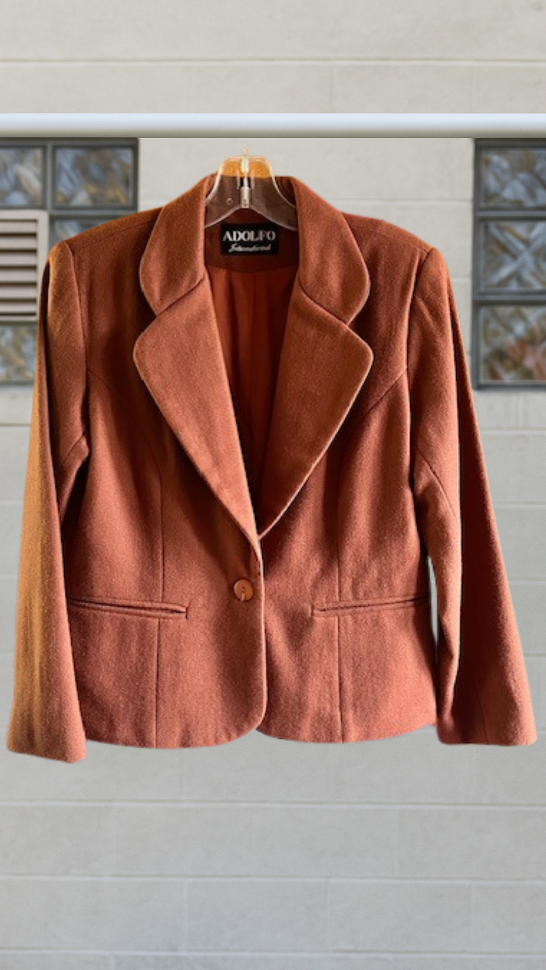 A cropped wool blazer in a warm oxblood-red tone with a notched lapel and single button closure, displayed against a neutral wall.
