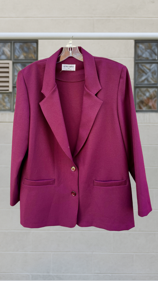 Oversized garnet blazer with strong shoulders, two-button front, and bold lapels, hanging on a hanger against a neutral block wall.
