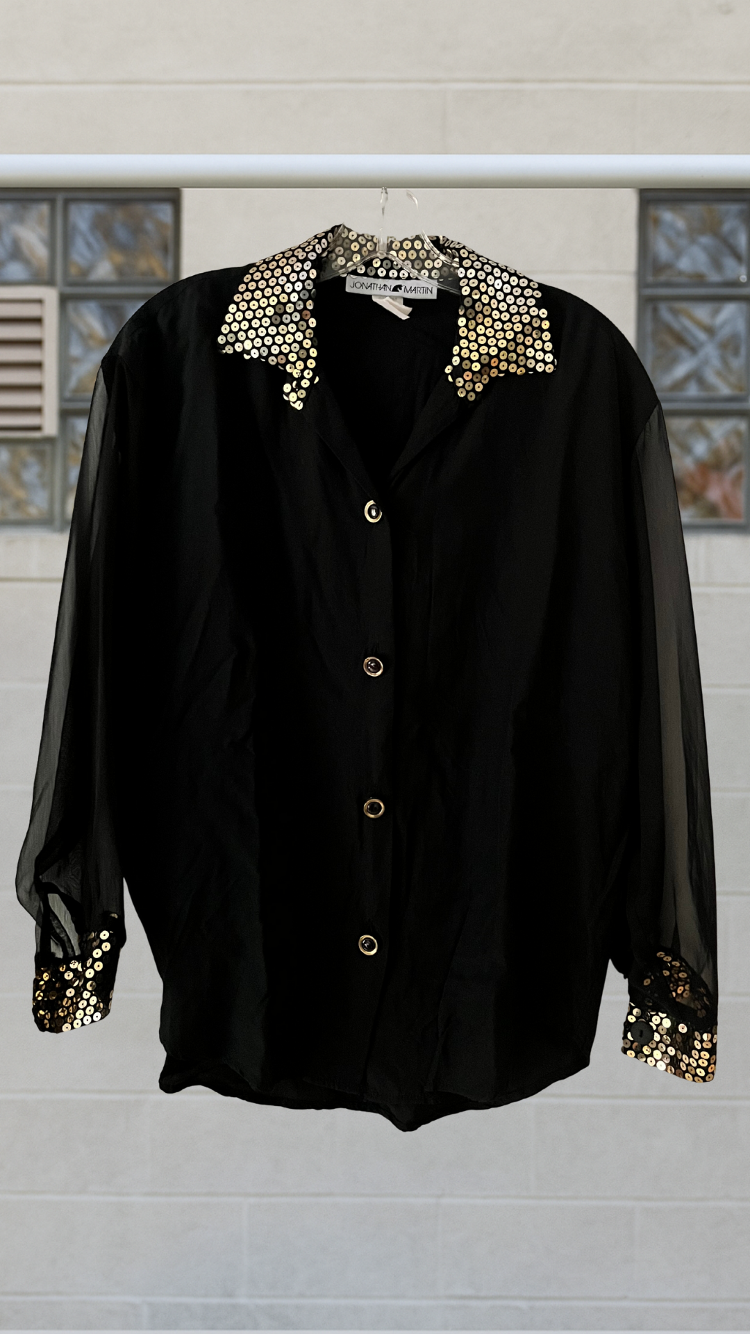 Black sheer blouse with gold sequined collar and cuffs, sheer sleeves, and button front, displayed on hanger outdoors.
