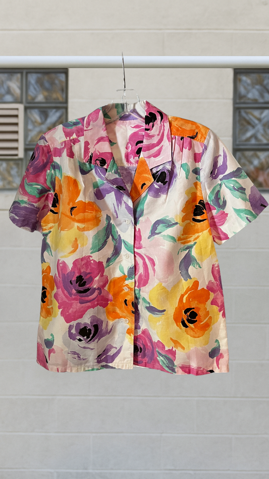 Short sleeve floral button-up shirt with vibrant painted blooms in orange, pink, purple, and yellow, hanging on a hanger outdoors.