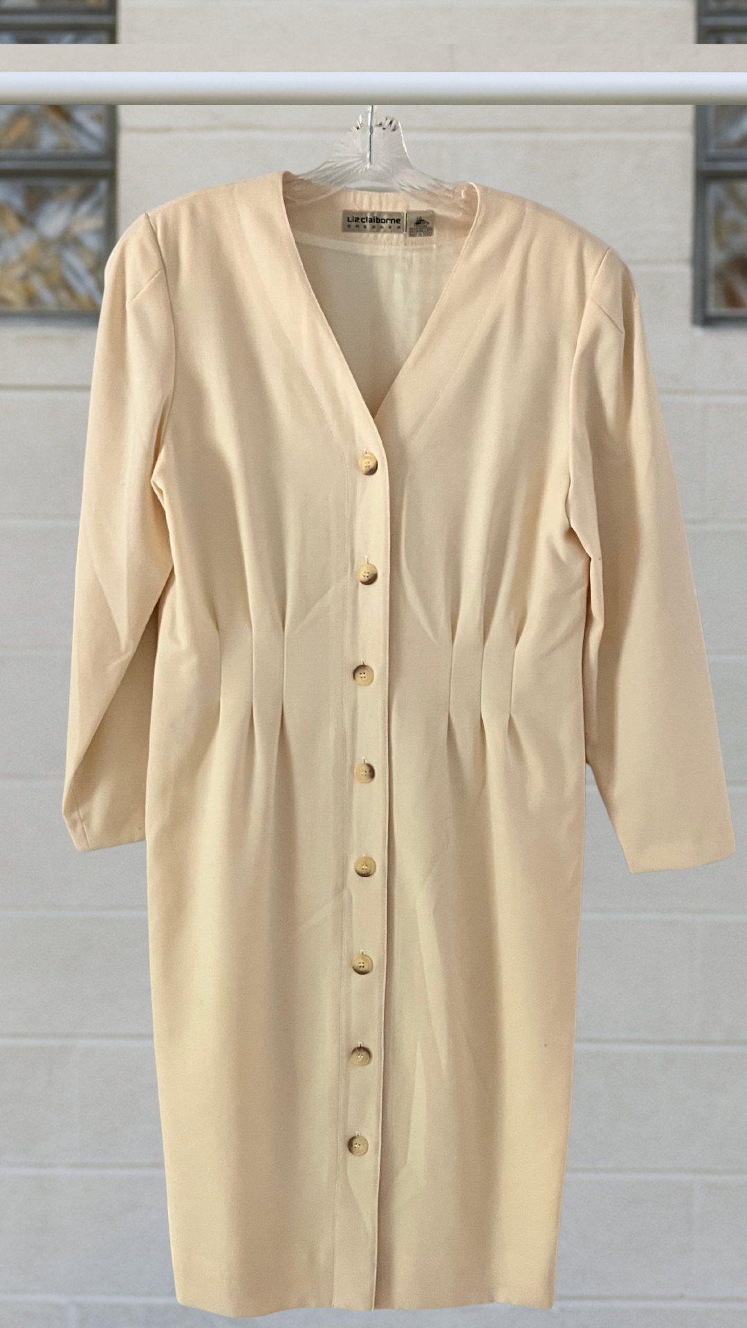 Ivory wool dress with V-neck, shoulder pads, pleated waist, long sleeves, and eight front buttons; hanging on a hanger against a light block-glass wall.
