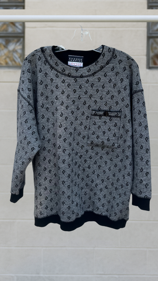 Grey paisley jacquard pullover with 3/4 sleeves, ribbed cuffs and hem, and a button-flap chest pocket; hanging on a hanger against a light block-glass wall.
