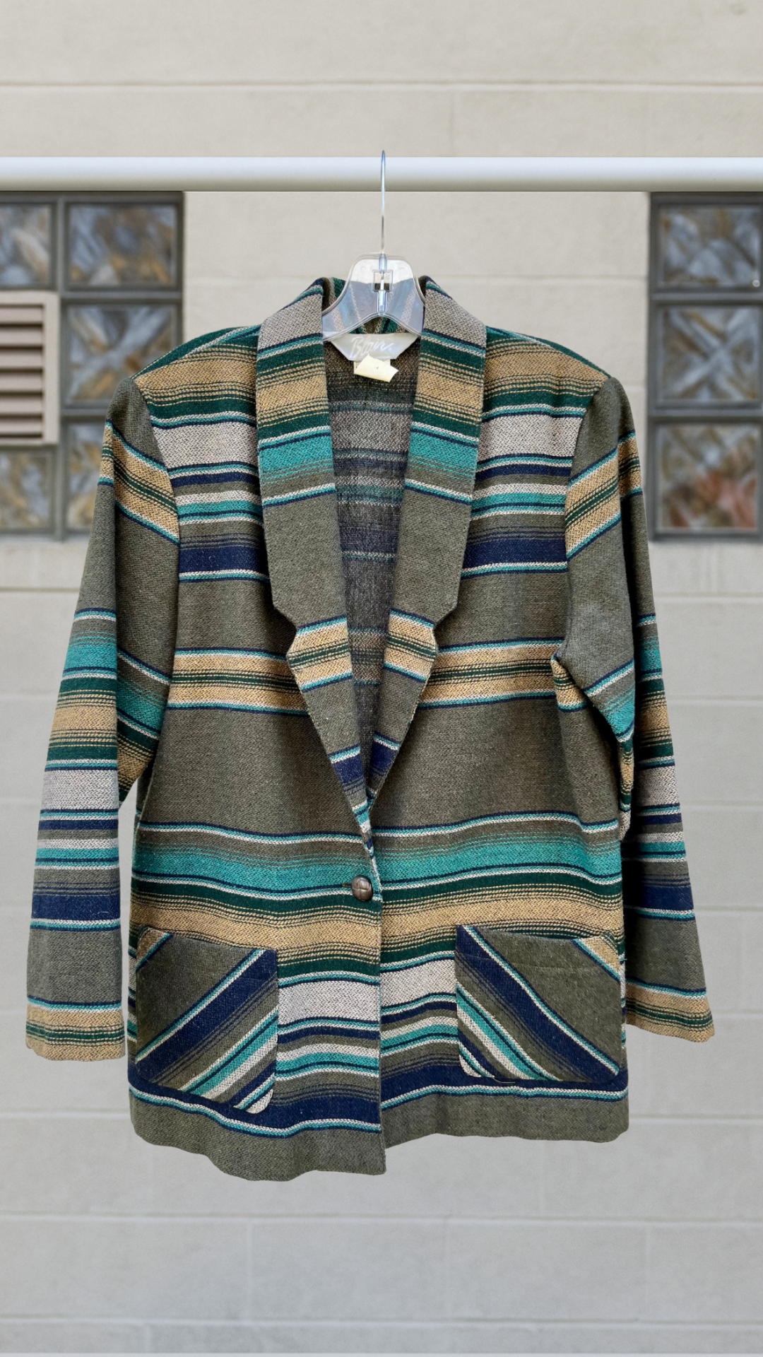 Full front view of a vintage striped blazer in earth tones and turquoise accents, hung on a hanger against a stone-textured wall.