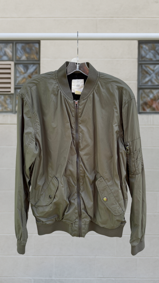 Olive drab bomber jacket with front zip and flap pockets, hanging against light gray tile wall