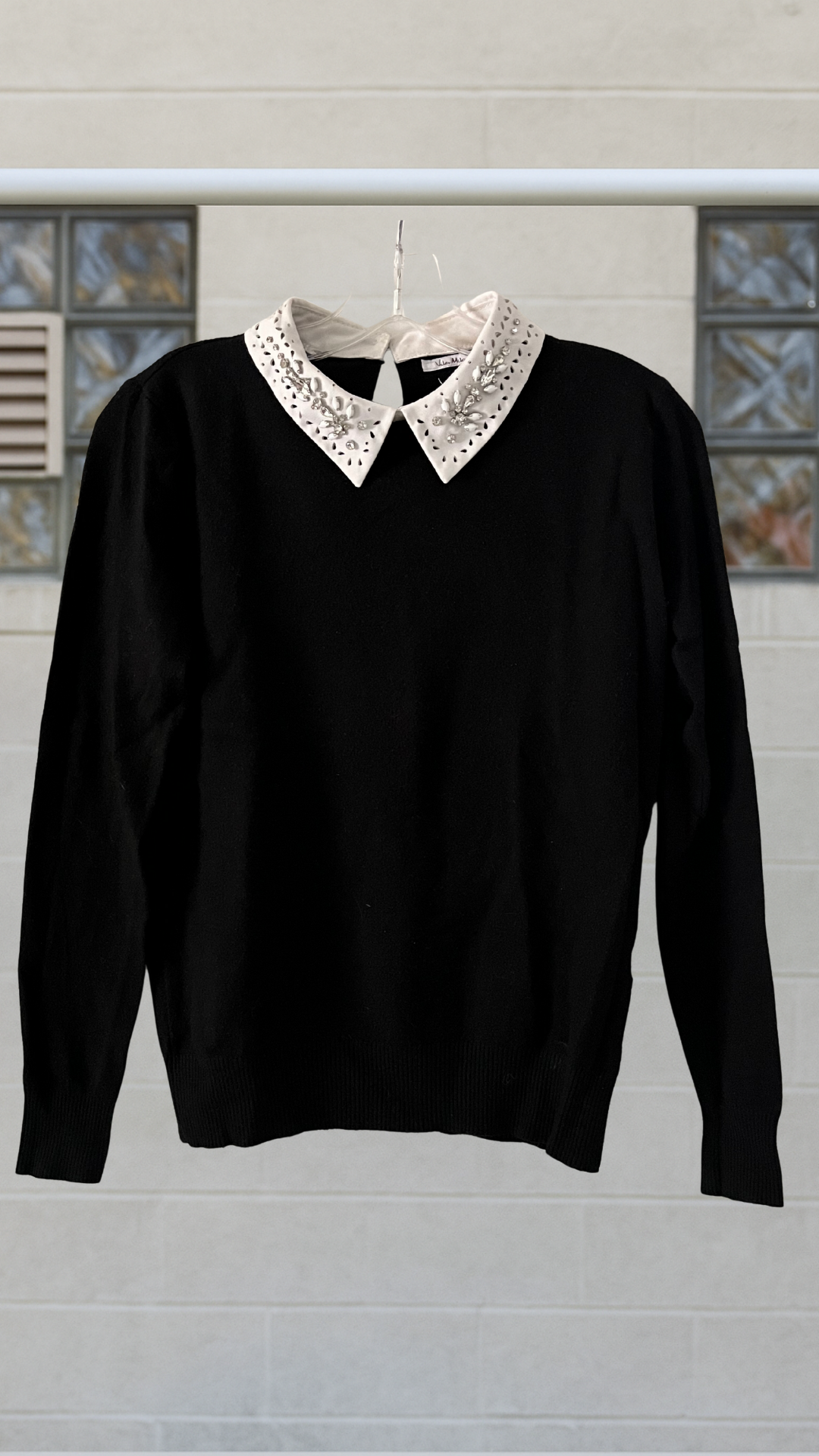 Black long-sleeve sweater with structured shape and a luminous white jeweled collar, hanging neatly against a neutral block wall.
