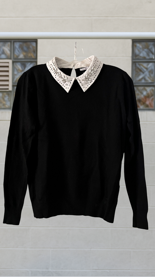 Black long-sleeve sweater with structured shape and a luminous white jeweled collar, hanging neatly against a neutral block wall.
