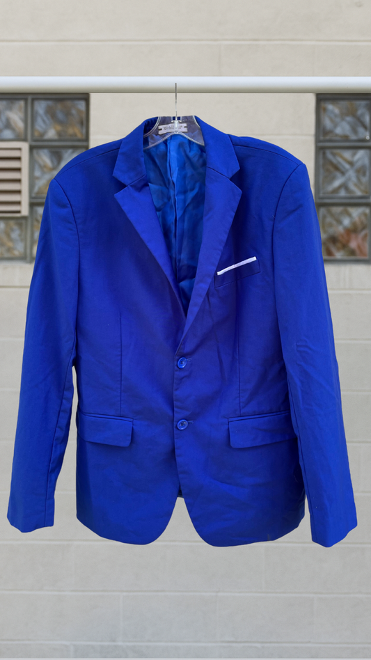 Bright cobalt blue blazer with notched lapels, two-button front, and white accent pocket, hanging on a hanger against a light brick wall.
