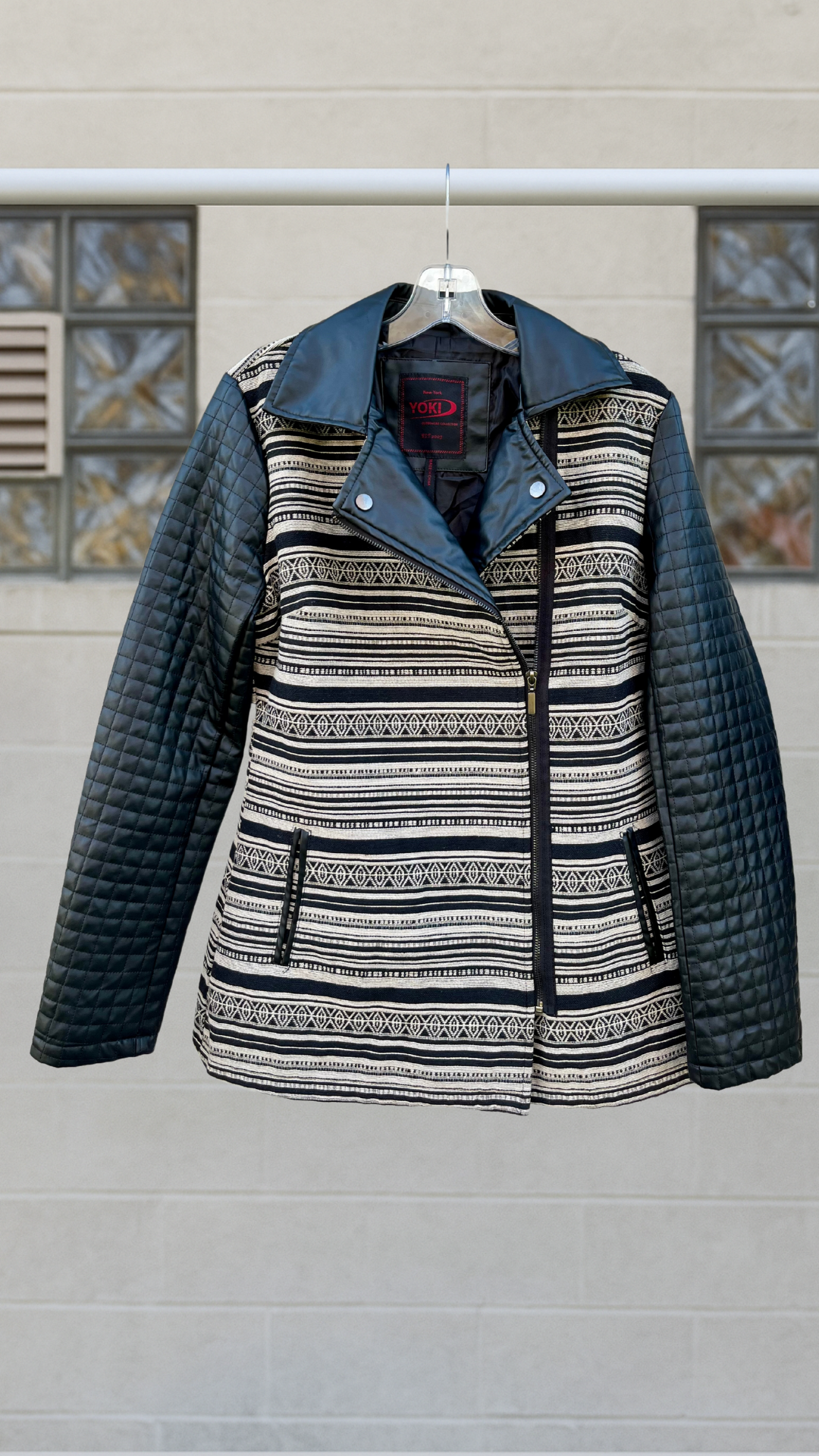Black and cream jacket with tribal-inspired horizontal pattern, quilted faux leather sleeves, and an asymmetrical zip front. Faux leather collar and snap lapels. Hanging on a clear hanger in front of a neutral background.