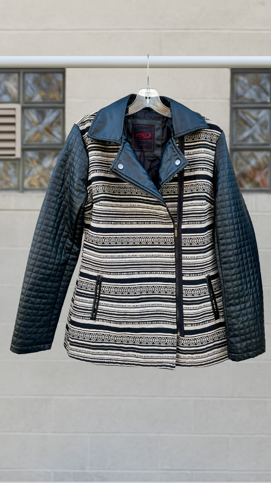 Black and cream jacket with tribal-inspired horizontal pattern, quilted faux leather sleeves, and an asymmetrical zip front. Faux leather collar and snap lapels. Hanging on a clear hanger in front of a neutral background.