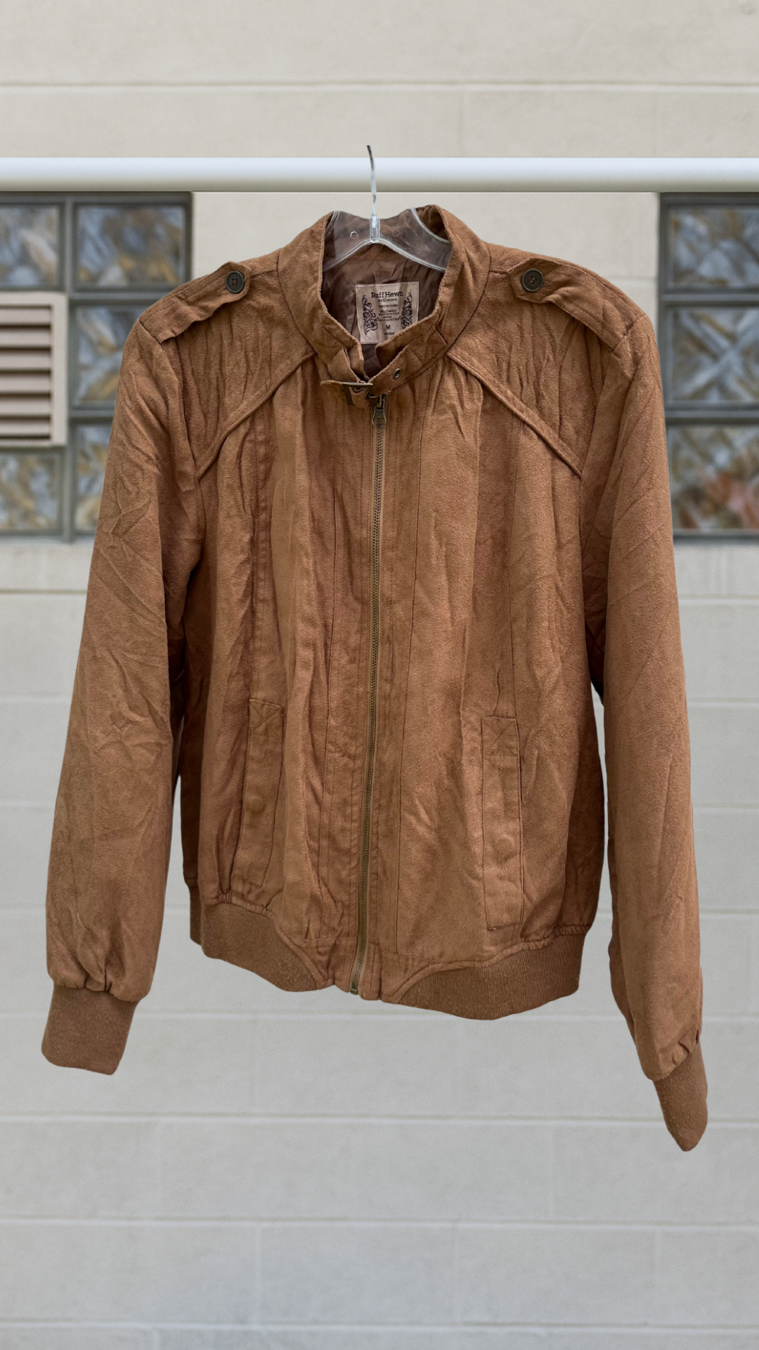 Light brown suede-like bomber jacket with zip front, mock neck strap, and epaulets, hanging on white wall background.