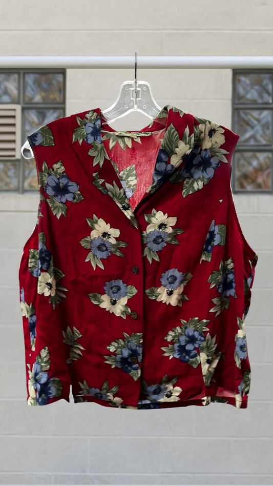 Sleeveless crimson rayon shirt with multi-color floral print, pointed collar, and wooden buttons, displayed front-on against light brick wall.
