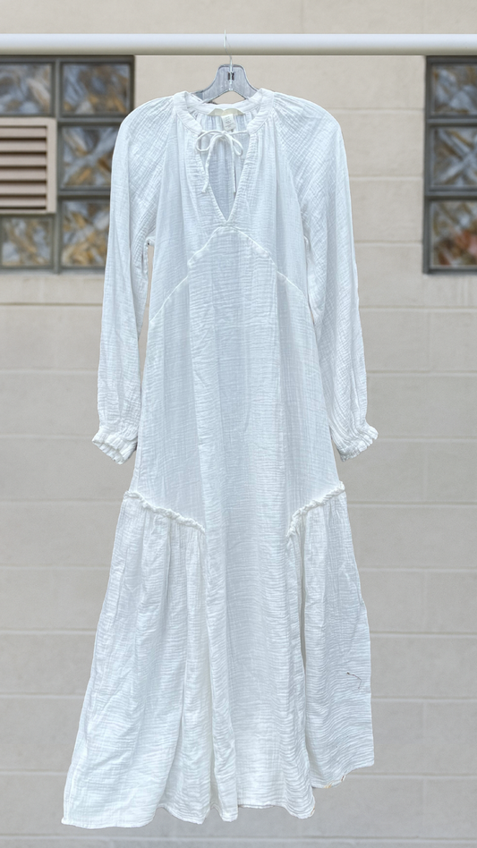 A long white cotton maxi dress with tie-neck detail, empire waist, and flowing full sleeves, hanging against a neutral block wall.