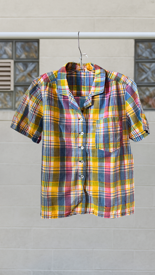 Short sleeve multicolor plaid button-up shirt with one front pocket, hanging outdoors on a hanger.