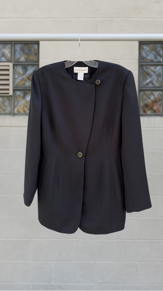 Vintage asymmetrical black blazer by Casual Corner, front view on hanger against block wall, subtle sheen and minimalist cut visible.