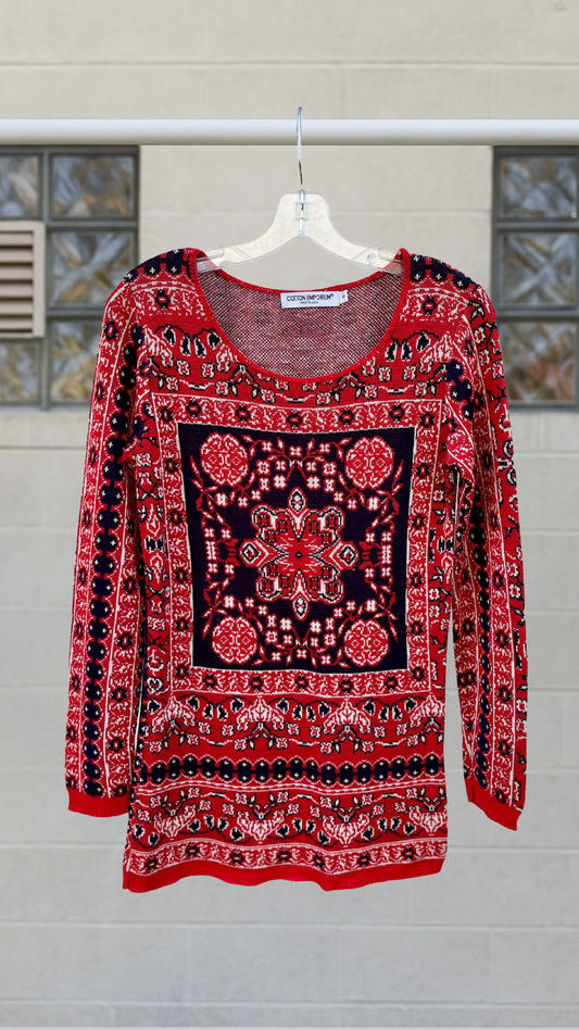 Red, navy, and white patterned long sleeve knit shirt with a central tapestry-inspired design, displayed on a clear hanger against a neutral background. Scoop neckline with thread detailing.