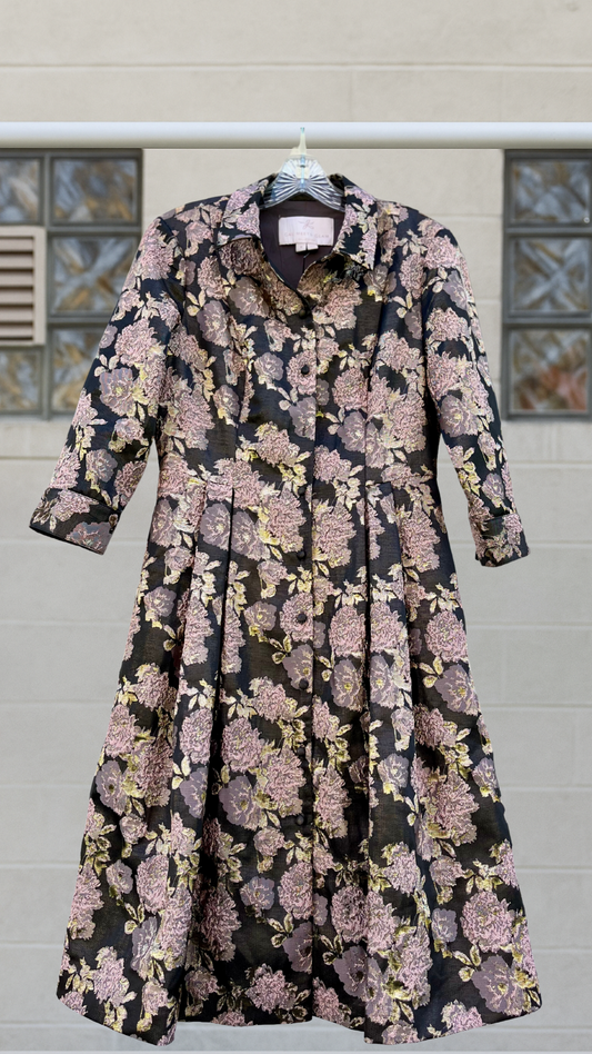 Vintage floral brocade coat-dress in black and gold with pink metallic roses, pleated skirt, and mid-length sleeves, hanging against a soft industrial background.