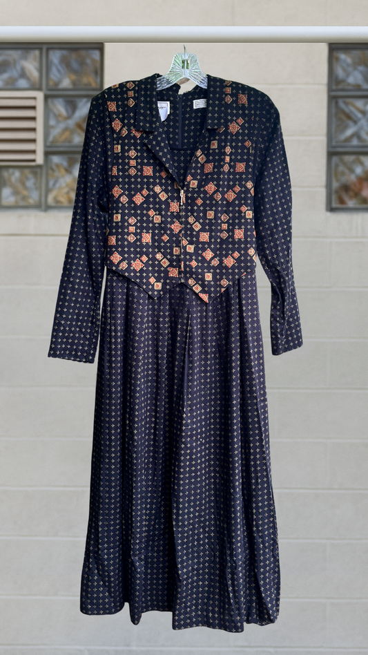 Full-length black rayon dress with rust and gold patterned vest overlay, long sleeves and pleated skirt, hanging against a pale brick wall.
