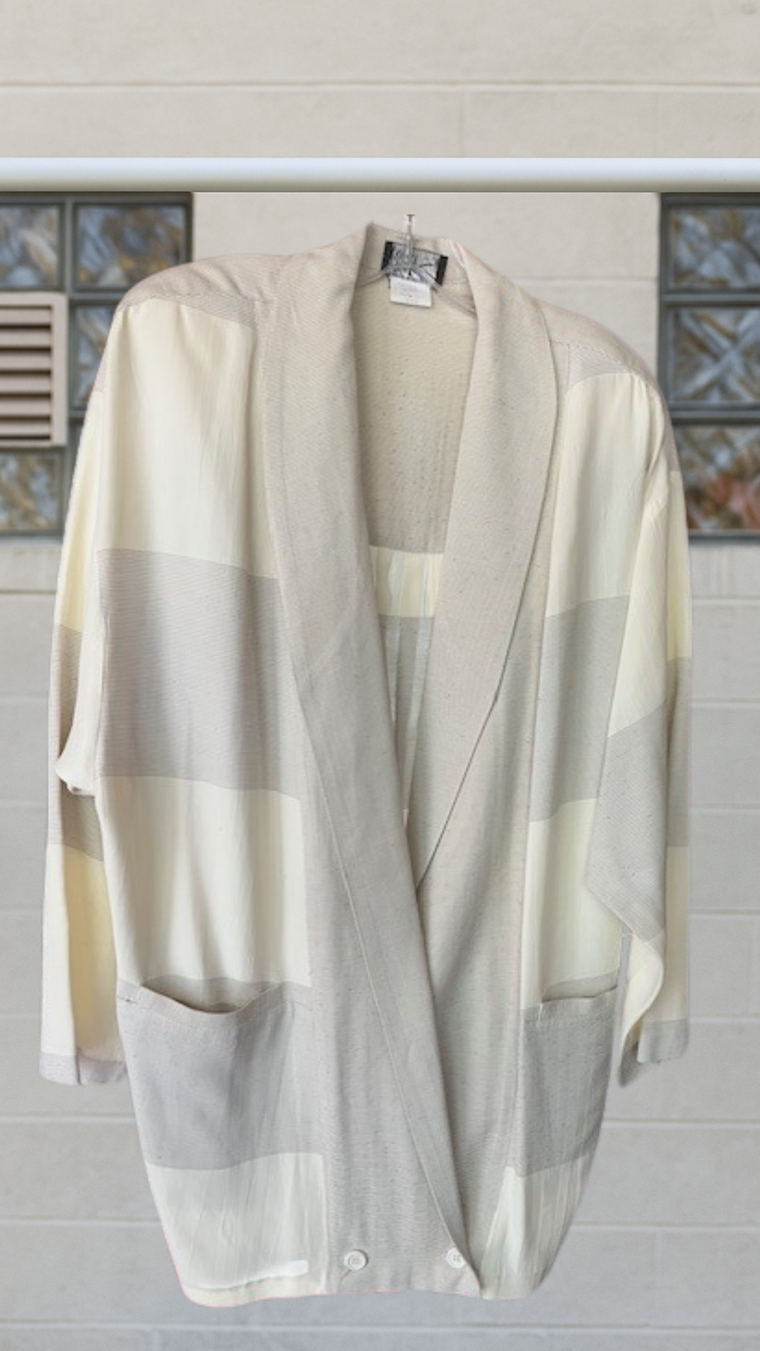 Front view of a softly structured vintage jacket with wide horizontal stripe-blocking in muted cream and grey tones, hanging on a hanger against a tiled wall.